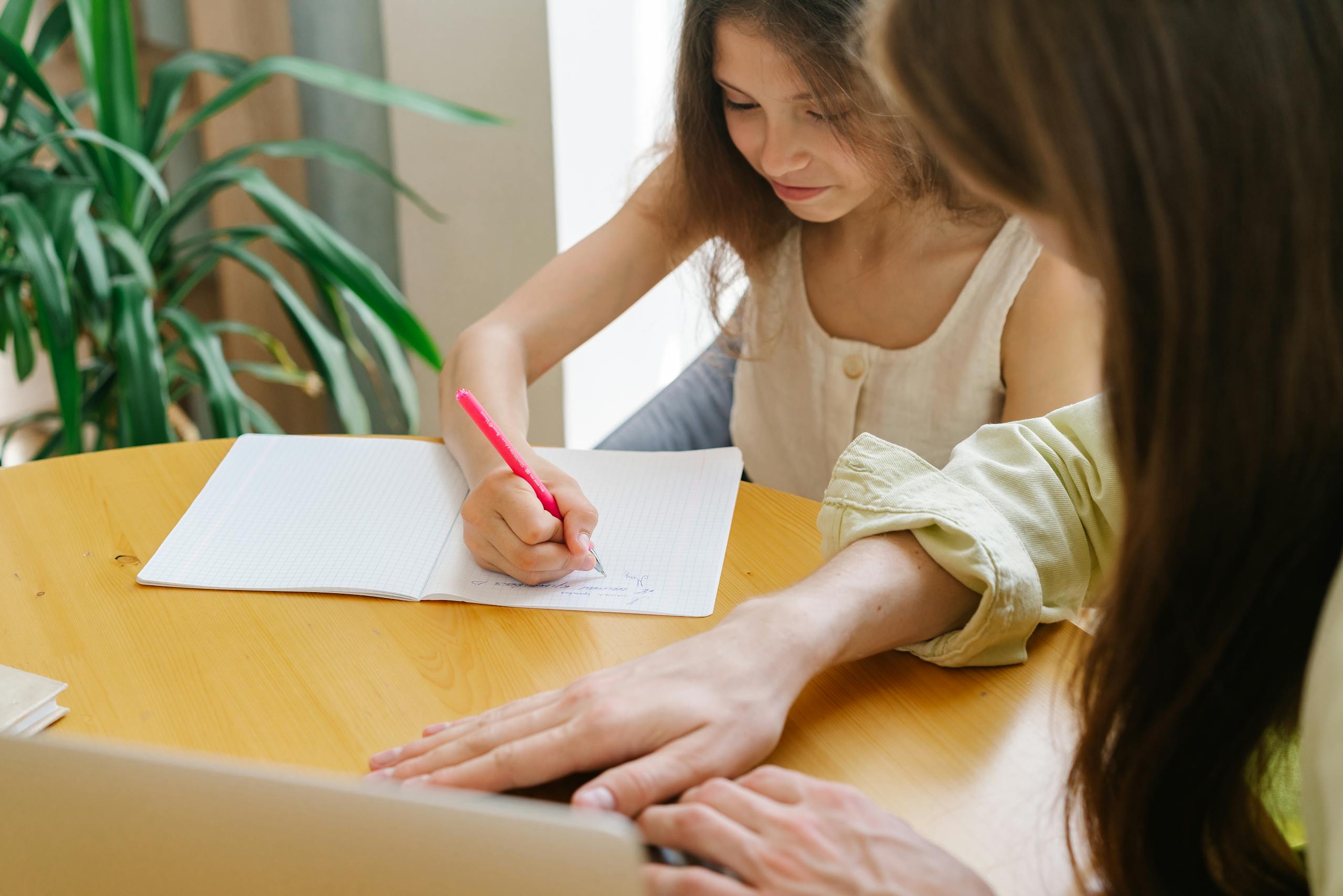 A woman helping a young girl with a writing task at a wooden table, enhancing their learning experience.